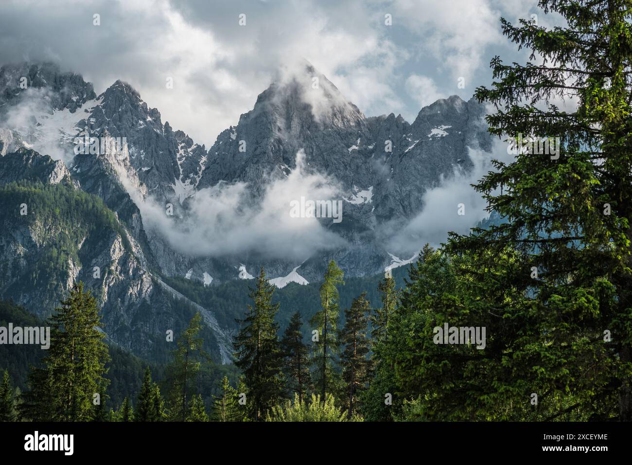 A scenic view of a mountain range in Slovenia, with snow-capped peaks and a thick layer of clouds swirling around them. The foreground features a dens Stock Photo