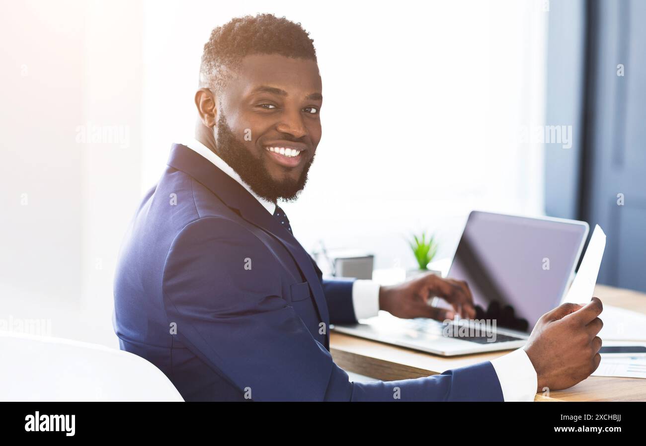 Cheerful black general manager checking reports, sitting in office Stock Photo