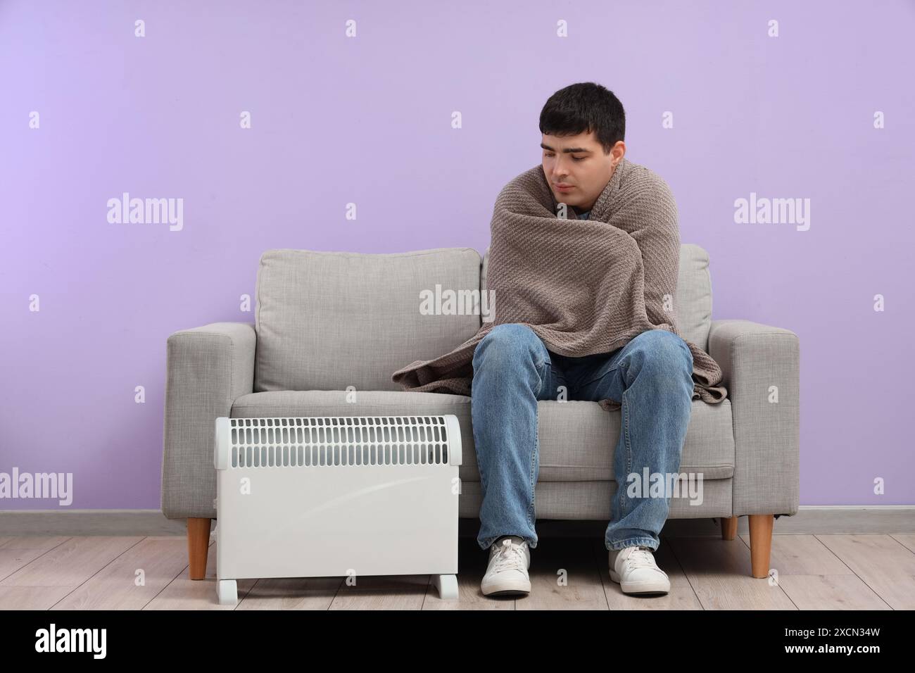 Frozen young man with plaid and radiator sitting on sofa near lilac ...