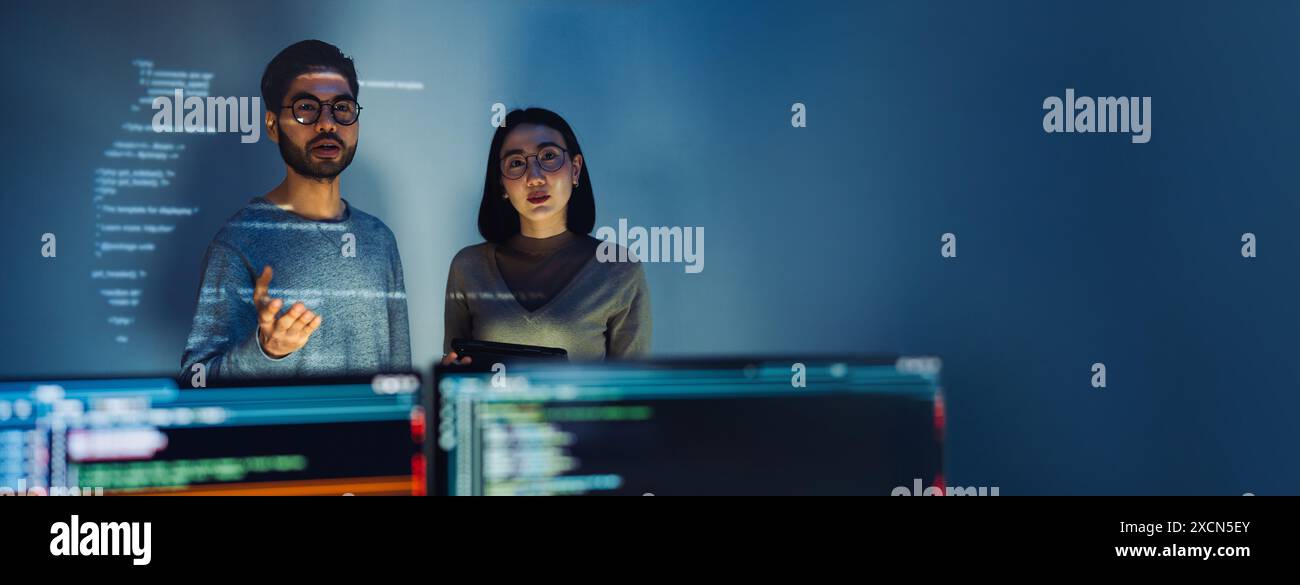 Asian Two software developers engaged in collaborative coding session, with code projected onto the wall and multiple computer screens displaying Stock Photo
