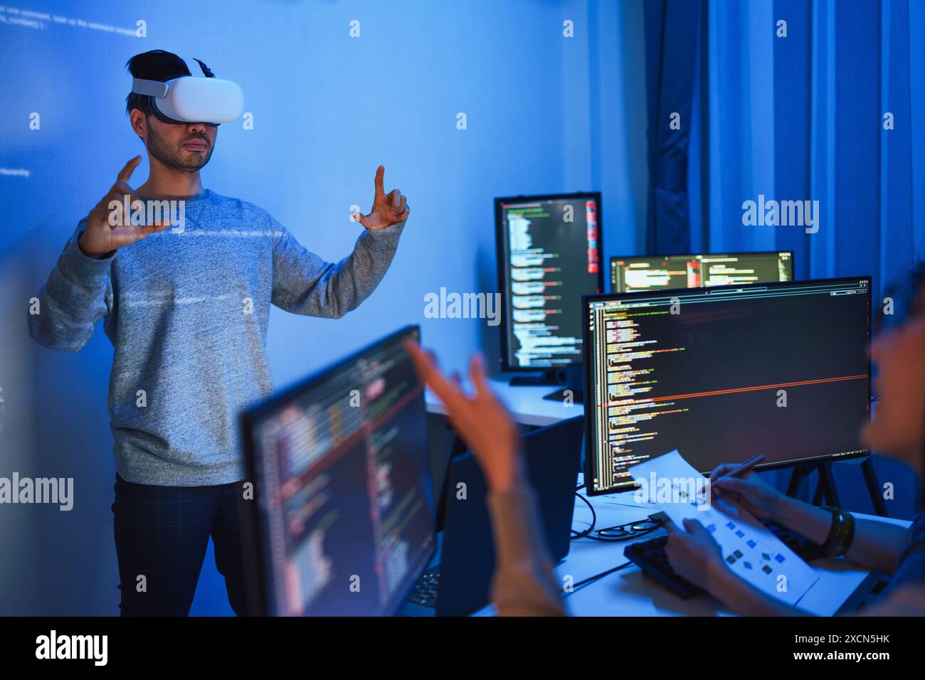 developer uses a VR headset to interact with projected interface designs, while colleagues discuss and code on screens, highlighting the integration o Stock Photo