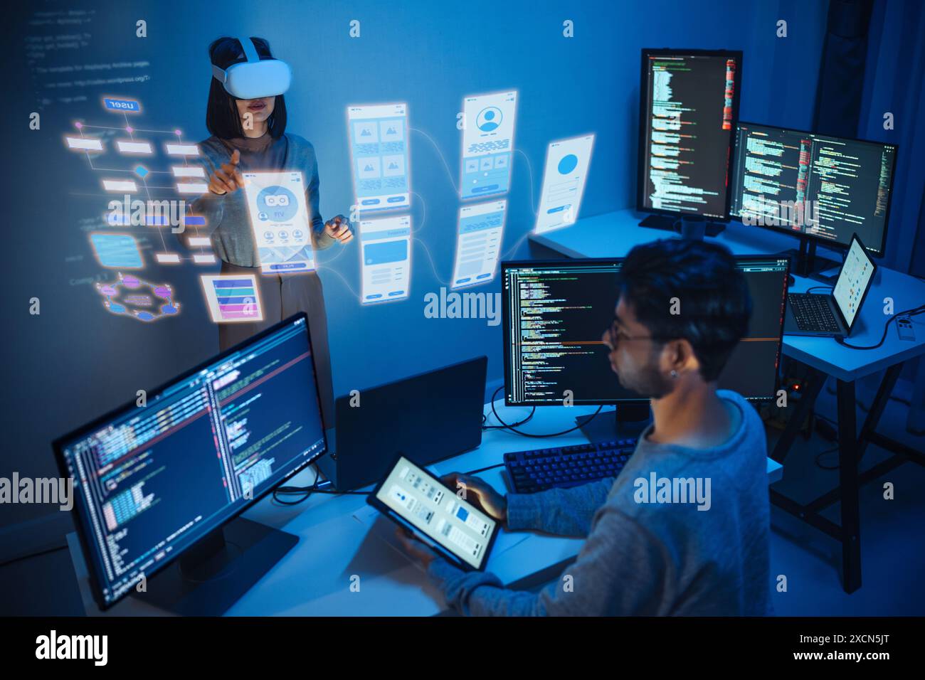 developer uses a VR headset to interact with projected interface designs, while colleagues discuss and code on screens at office Stock Photo