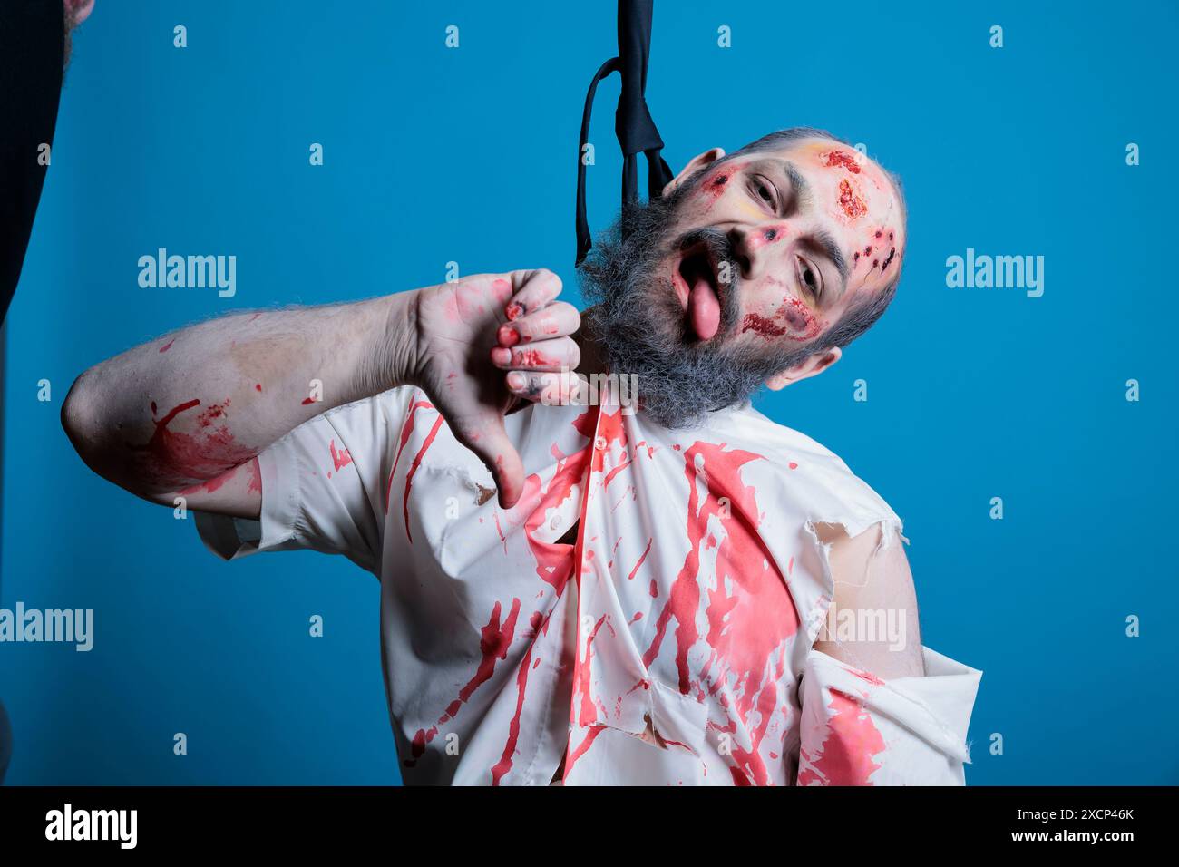 Unhappy man turned into zombie by overworking, committing suicide, feeling burdened by capitalism grind demands. Upset employee shows thumbs down sign, hanging himself with tie, studio background Stock Photo