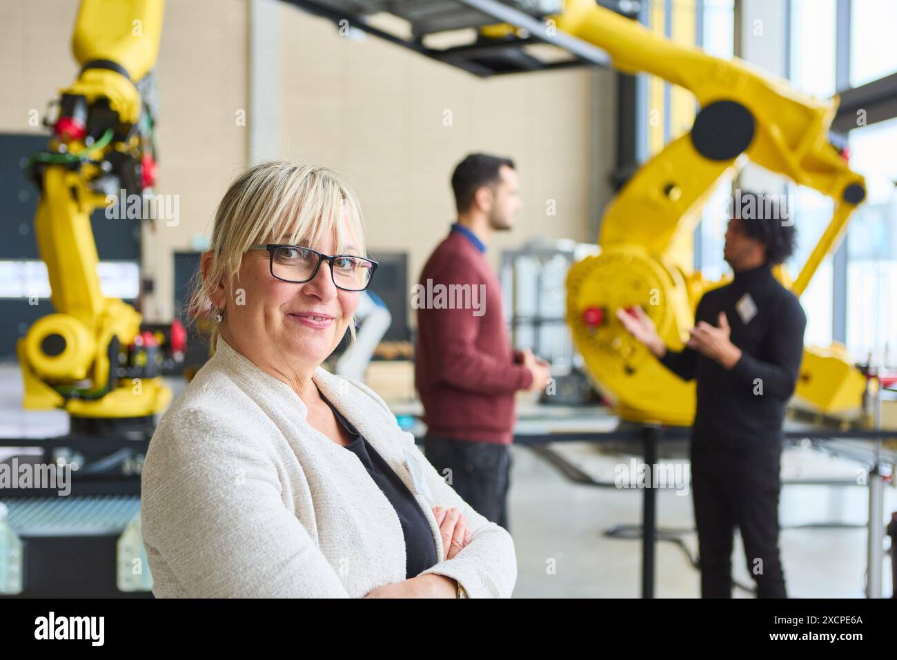 Confident woman standing in robotics lab while colleagues discuss automation. Focus on teamwork, robotics training, and industrial technology. Stock Photo