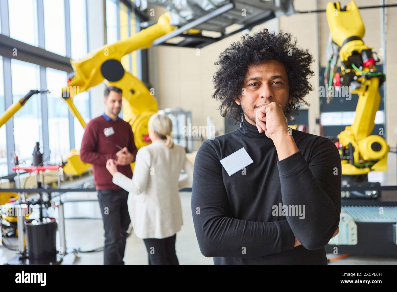 Confident engineer poses in robotics lab while colleagues discuss robot development, showcasing industrial automation and innovation. Stock Photo