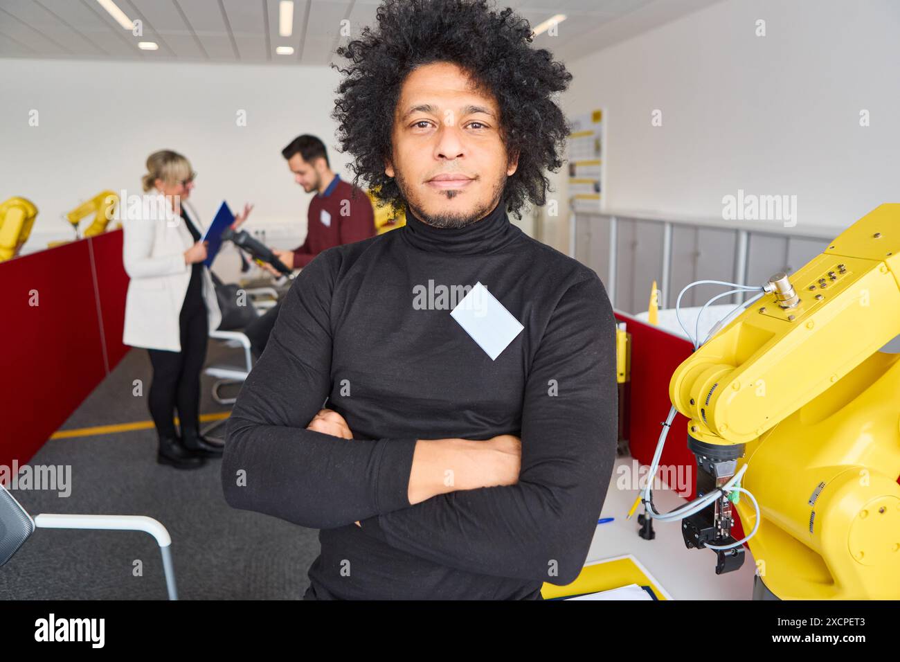Engineer standing confidently in a robotics lab with students and robotic arms, showcasing modern technology and teamwork. Stock Photo