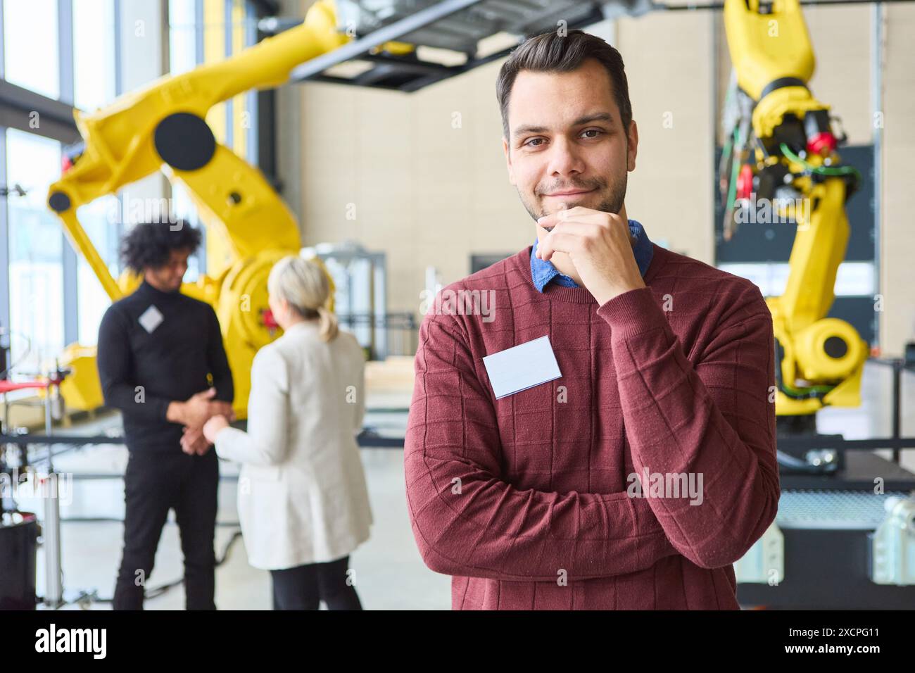 Confident engineer standing in a robotics lab with coworkers discussing automation projects near industrial robots equipped with gripper arms Stock Photo