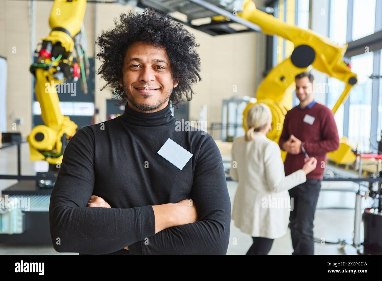 Confident engineer standing in a robotics workshop with colleagues discussing robotic arm programming and industrial automation in the background. Stock Photo