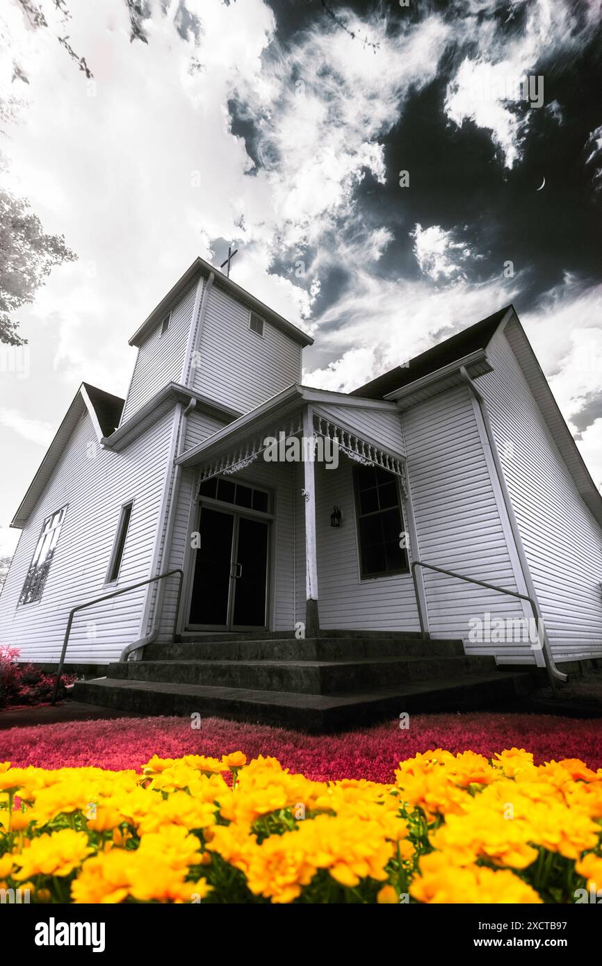 A small rural church beneath a muted blue sky, crescent moon. Red grass and golden flowers are below. Cross on belfry appears in cloud opening. Stock Photo