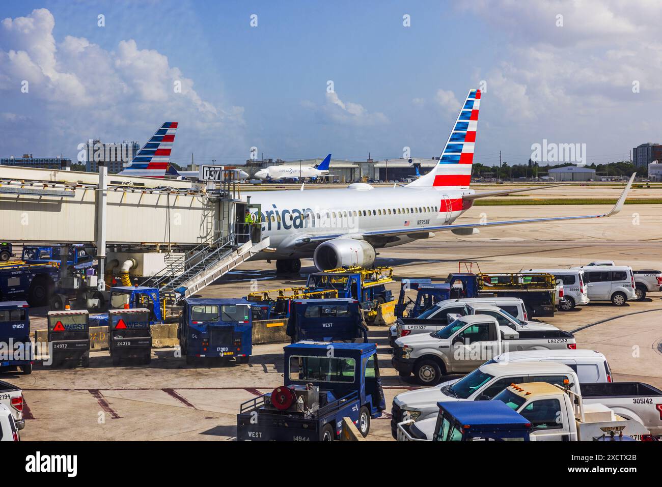 View of Miami airport runway with cargo trucks and American Airlines plane connected to jet bridge for passenger boarding. Miami. USA. Stock Photo