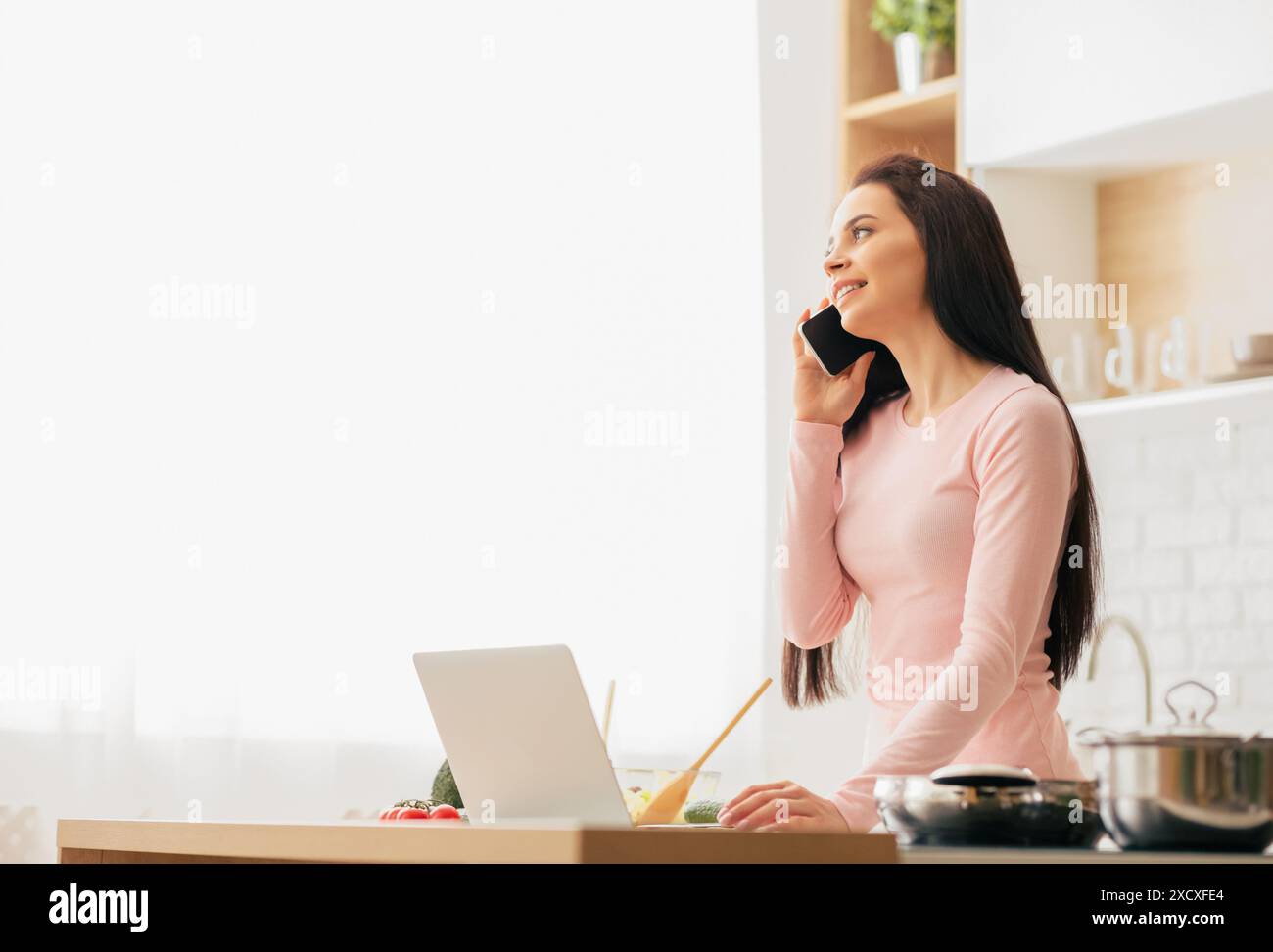 Woman Talking on Phone While Using Laptop, Kitchen Interior Stock Photo
