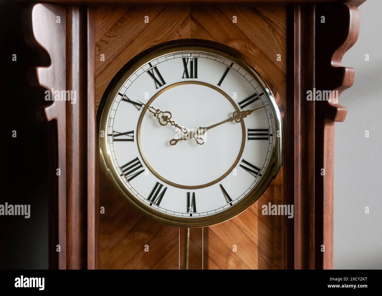 An antique wooden wall mounted grandfather clock with roman numerals and a glass door on a dimly
