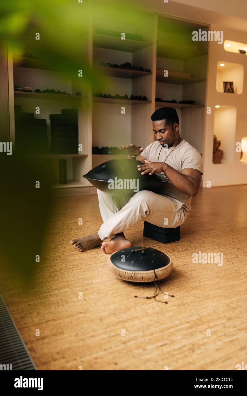 Vertical shot of African man playing on modern musical instrument Hand pan, Vadjraghanta, metal tongue drum sitting cross-legged on floor at home. Stock Photo