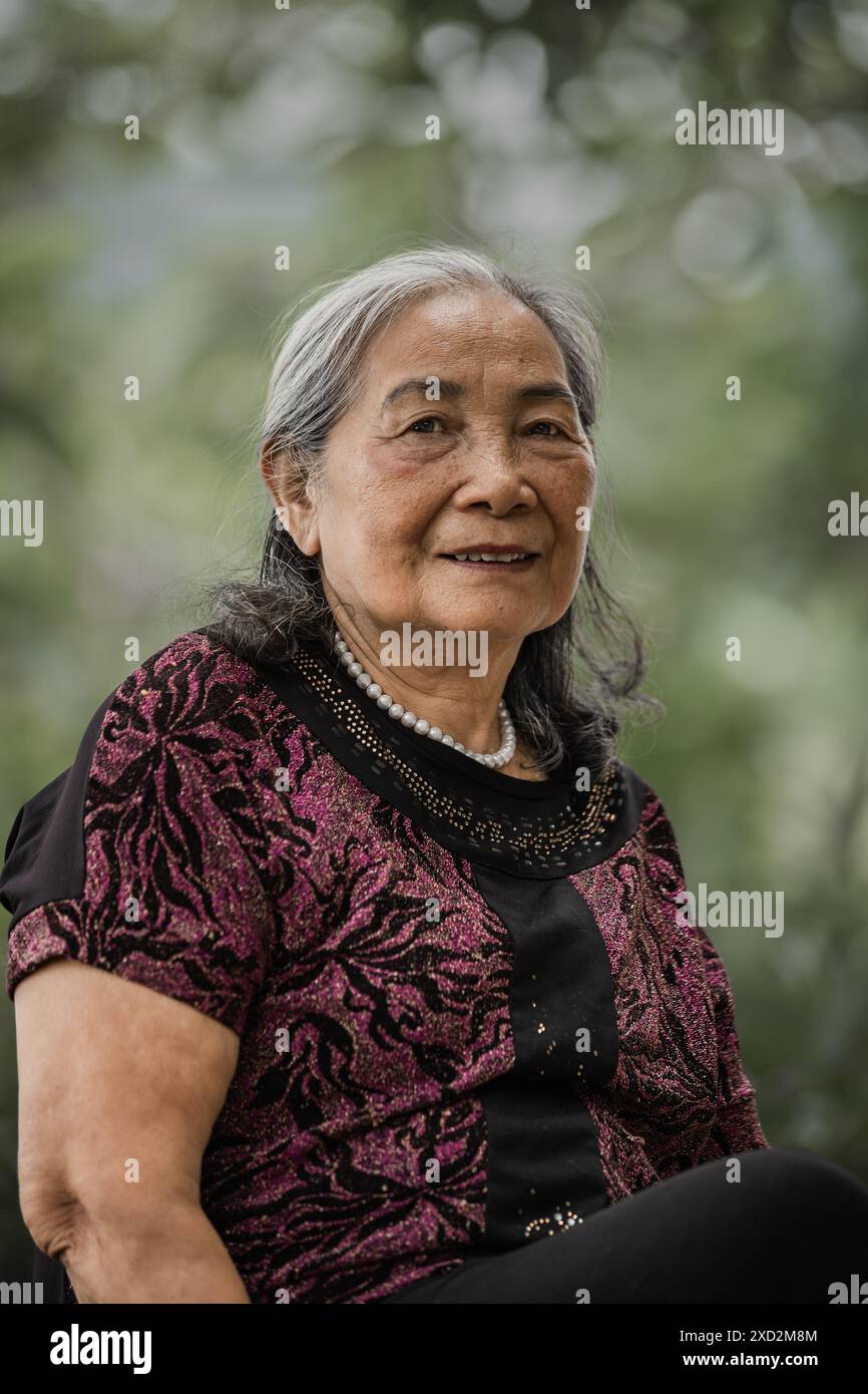 Portrait of an Elderly woman at the park. Asian Senior woman sitting on a bench in a summer park. Old smiling lady sits on a park bench in Vinh Minh V Stock Photo