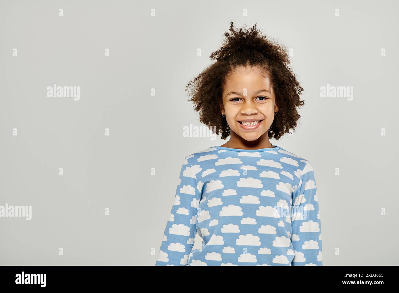 A young African American girl happily wearing a blue shirt with clouds pattern, posing on a grey background. Stock Photo