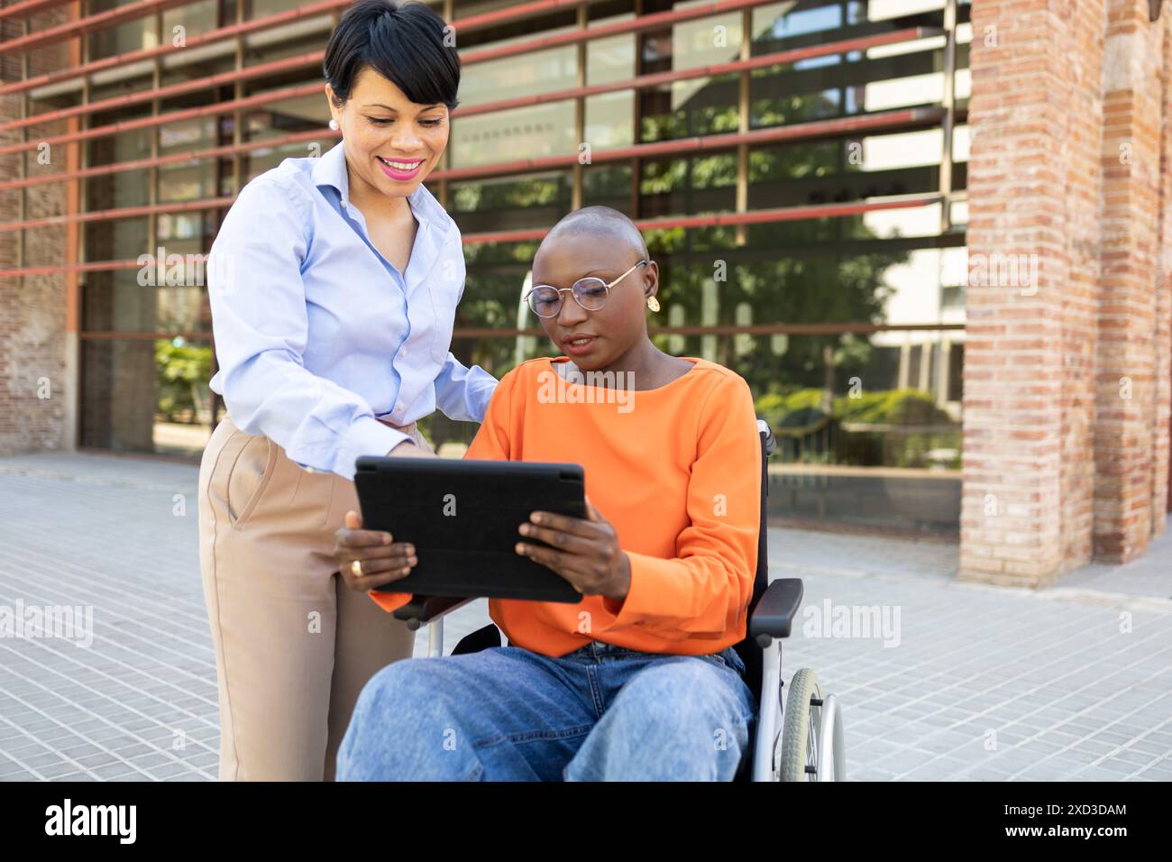 A professional Latin American woman assists a young African American woman in a wheelchair, using a digital tablet outdoors near a modern office build Stock Photo