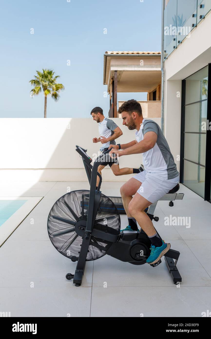 Two men focused on their workout using a stationary bike and treadmill in an outdoor home gym with a scenic palm view Stock Photo Alamy