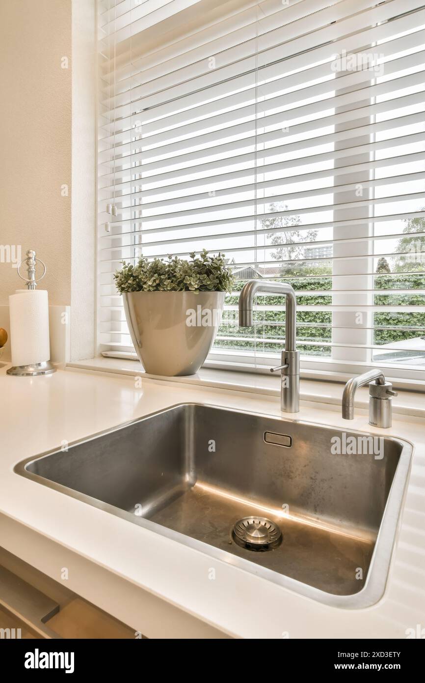 A kitchen sink in front of a window with blinds on the windowsilling it is white and there is a pot of flowers Stock Photo