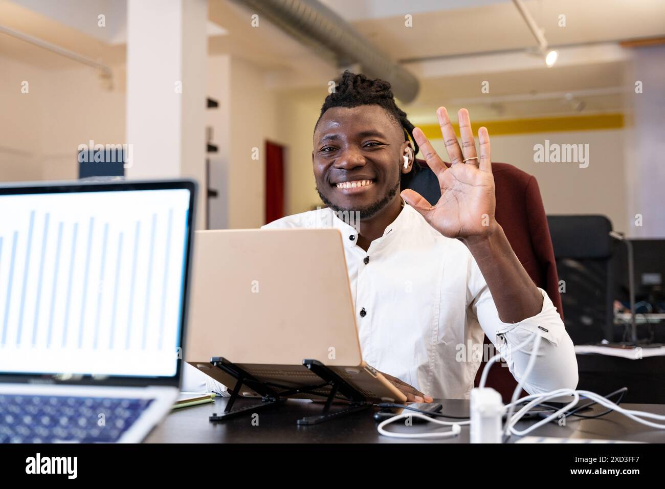 A dynamic African-American man in a modern office waves cheerfully at his laptop camera, signaling a friendly greeting or successful virtual meeting Stock Photo