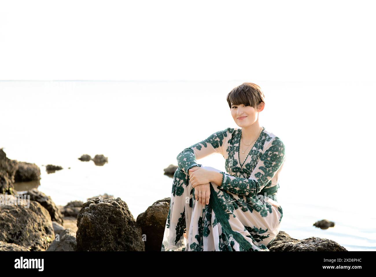 A woman in a patterned dress sitting on rocks by the water, smil Stock ...