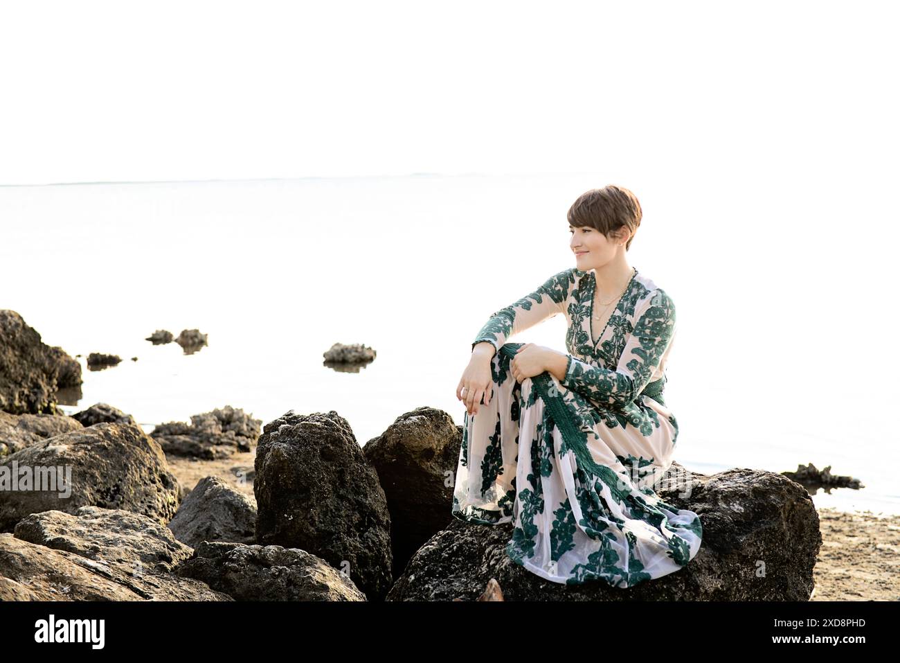 A woman in a patterned dress sitting on rocks by the water, smil Stock ...