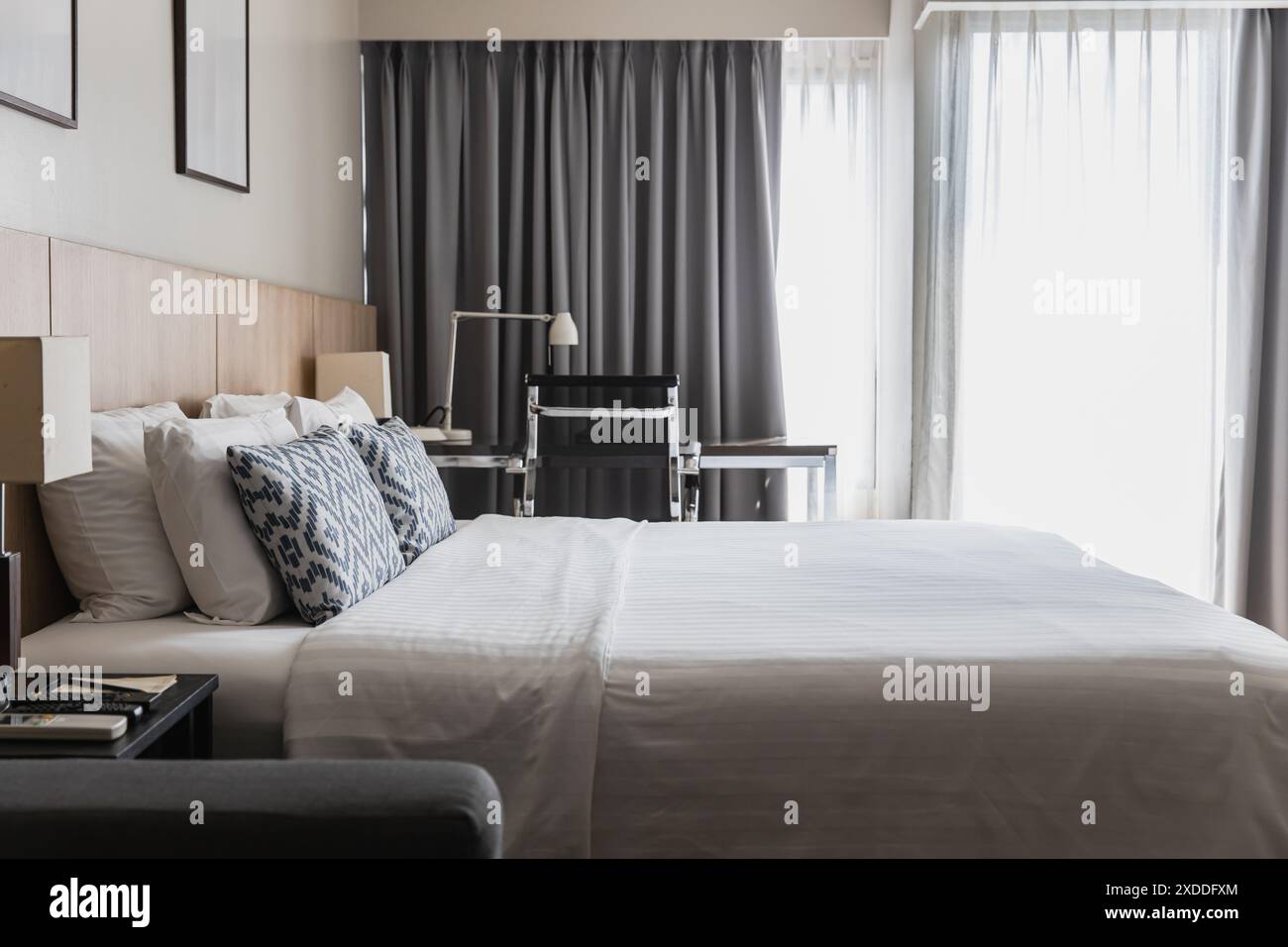 Modern bedroom featuring a neat bed, decorative pillows, a sleek desk, and large windows with curtains. Ideal for concepts of comfort, style, and cont Stock Photo