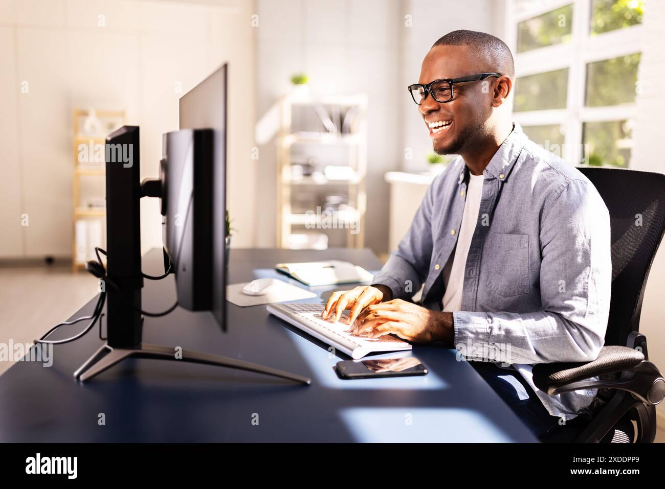 Efficient African American Businessman Managing Documents and Multiple Computer Screens at Corporate Office Desk, Smiling While Working. Stock Photo