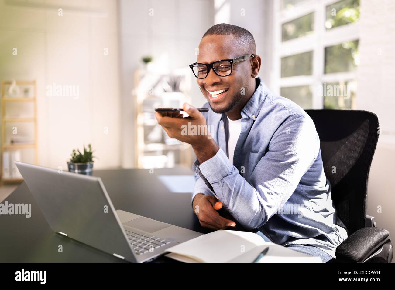 Professional African American Businessman Using Smartphone App for Client Call in Modern Office Setting with Laptop and Smiling Stock Photo