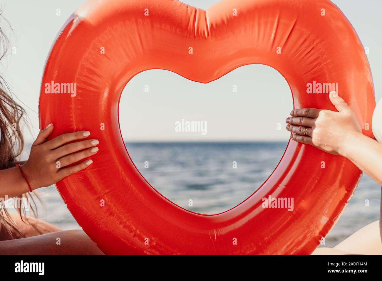 Two women are holding a red inflatable heart on the beach. The women