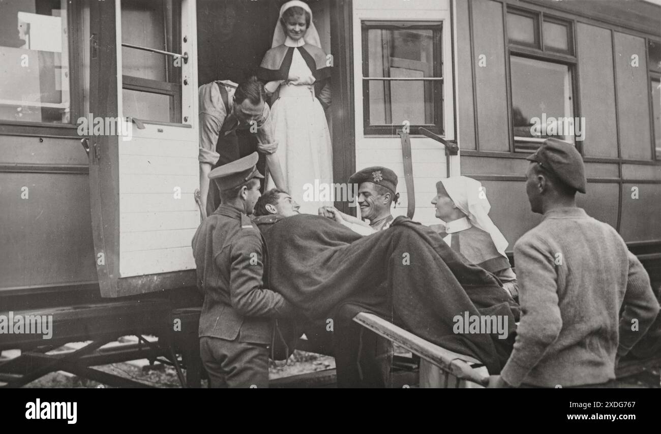 A black and white postcard depicting a wounded soldier being loaded onto a train. The soldier is lying on a stretcher, being carried by two men in uniform. Two other men in uniform stand on either side of the stretcher, while two nurses stand behind. The scene takes place at a train station, with the open doors of a train car visible in the background. The image likely portrays the transportation of a wounded British soldier back to England during World War I. Stock Photo