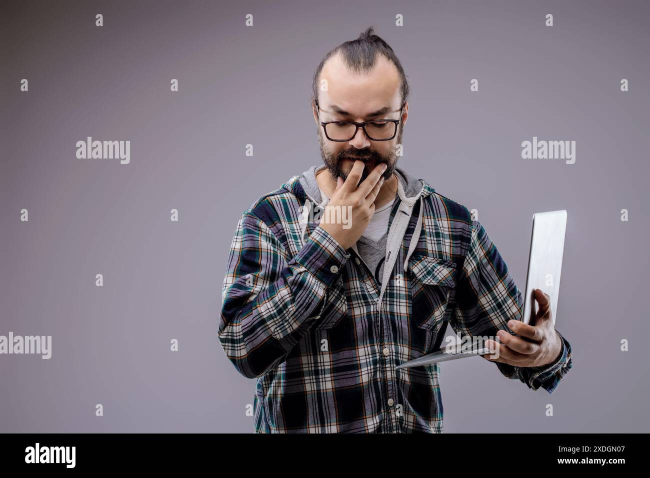 Focused web developer with laptop contemplates coding problem in studio. Gray background enhances serious atmosphere. Expertise and determination evid Stock Photo