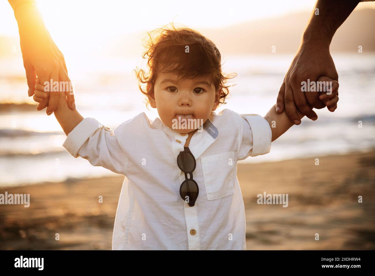 A one-year-old boy in a white shirt and sunglasses walks on the beach, holding his parents' hands. The background features sand and the warm light of Stock Photo