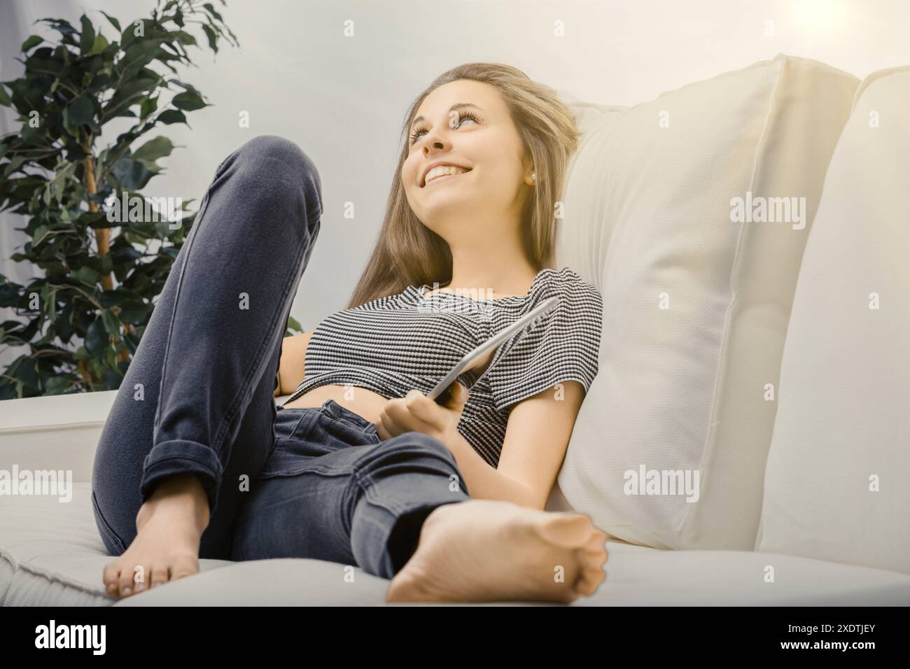 Young woman relaxes on her sofa with a tablet, smiling and daydreaming ...