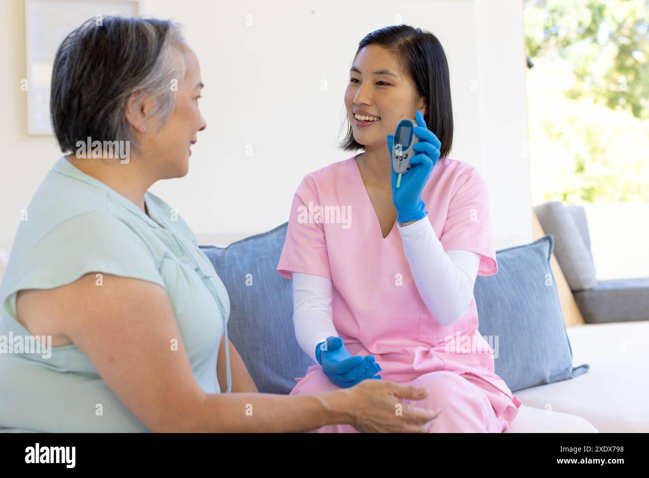 Female nurse in pink scrubs showing blood glucose meter to senior woman Stock Photo