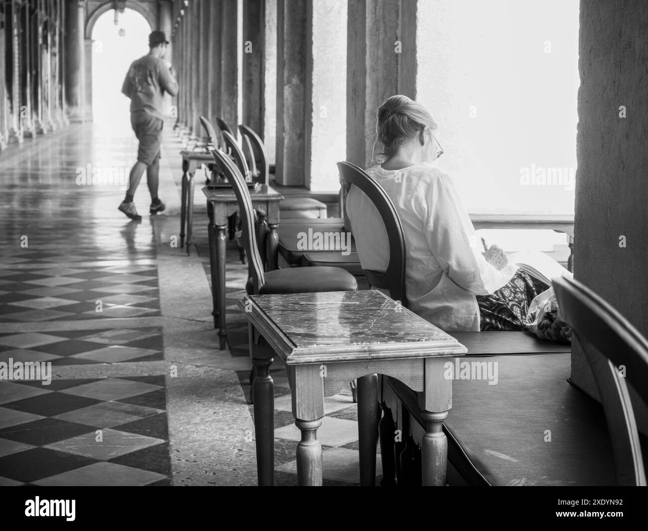 Venice, Italy - June 30th 20220 Black and white photo of a woman enjoying a book in a covered passageway in venice as a tourist walks in the backgroun Stock Photo