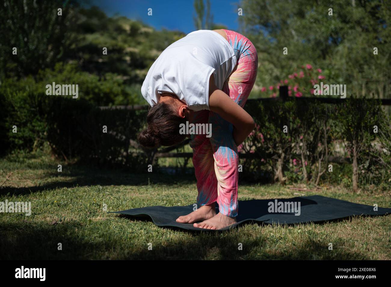 A woman performs a yoga forward bend on a mat in a sunny park ...