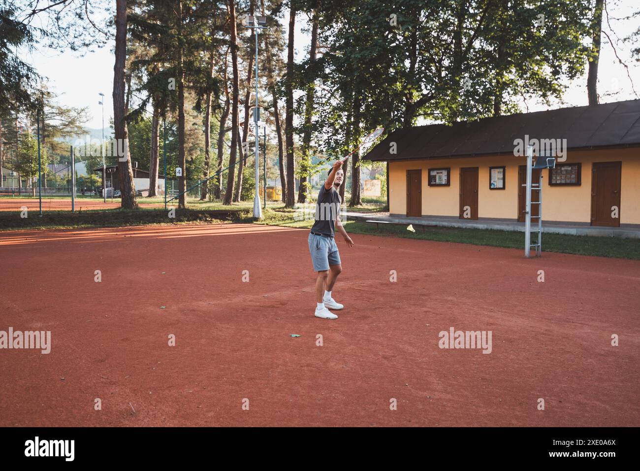 Black-haired athlete with a smile on his face plays badminton on a clay court. Enthusiasm for amateur sport. Learning new movement skills. Stock Photo