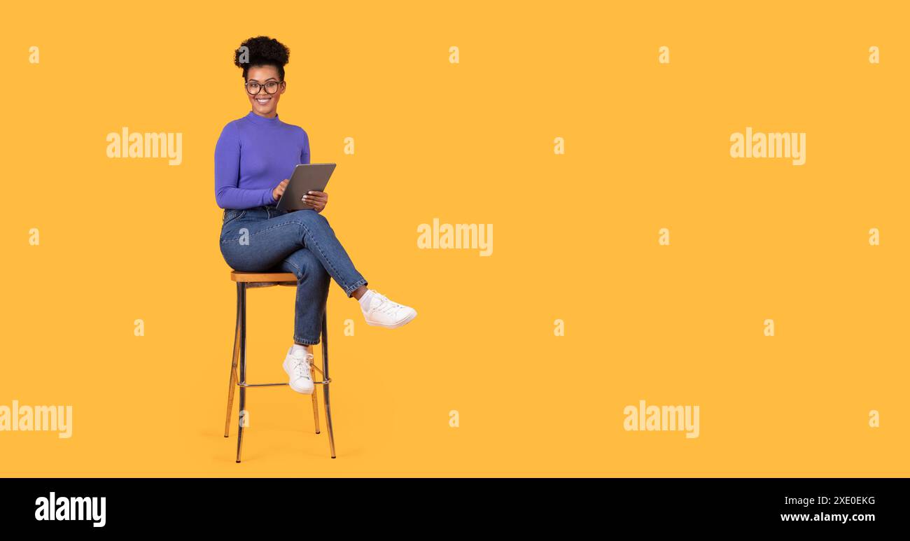 Smiling Woman Using Tablet While Sitting on Stool Against Yellow Stock Photo