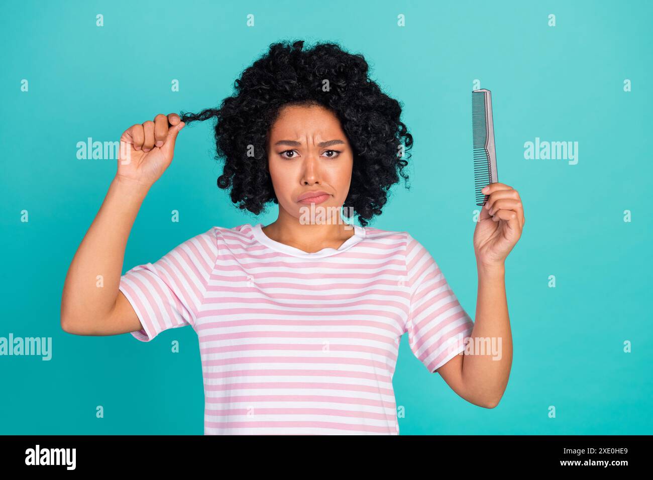 Photo of young beautiful lady in striped t shirt upset holding to untangle her hair using comb isolated on aquamarine color background Stock Photo