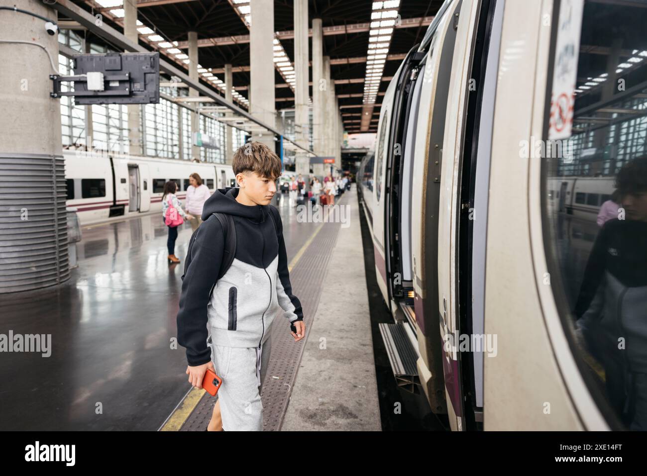 Teenager boarding in a high-speed train station, holding a phone and carrying a backpack. Stock Photo