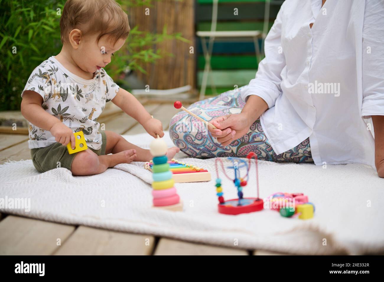 A mother and her baby play together on a blanket spread over a wooden deck, enjoying colorful educational toys. Stock Photo