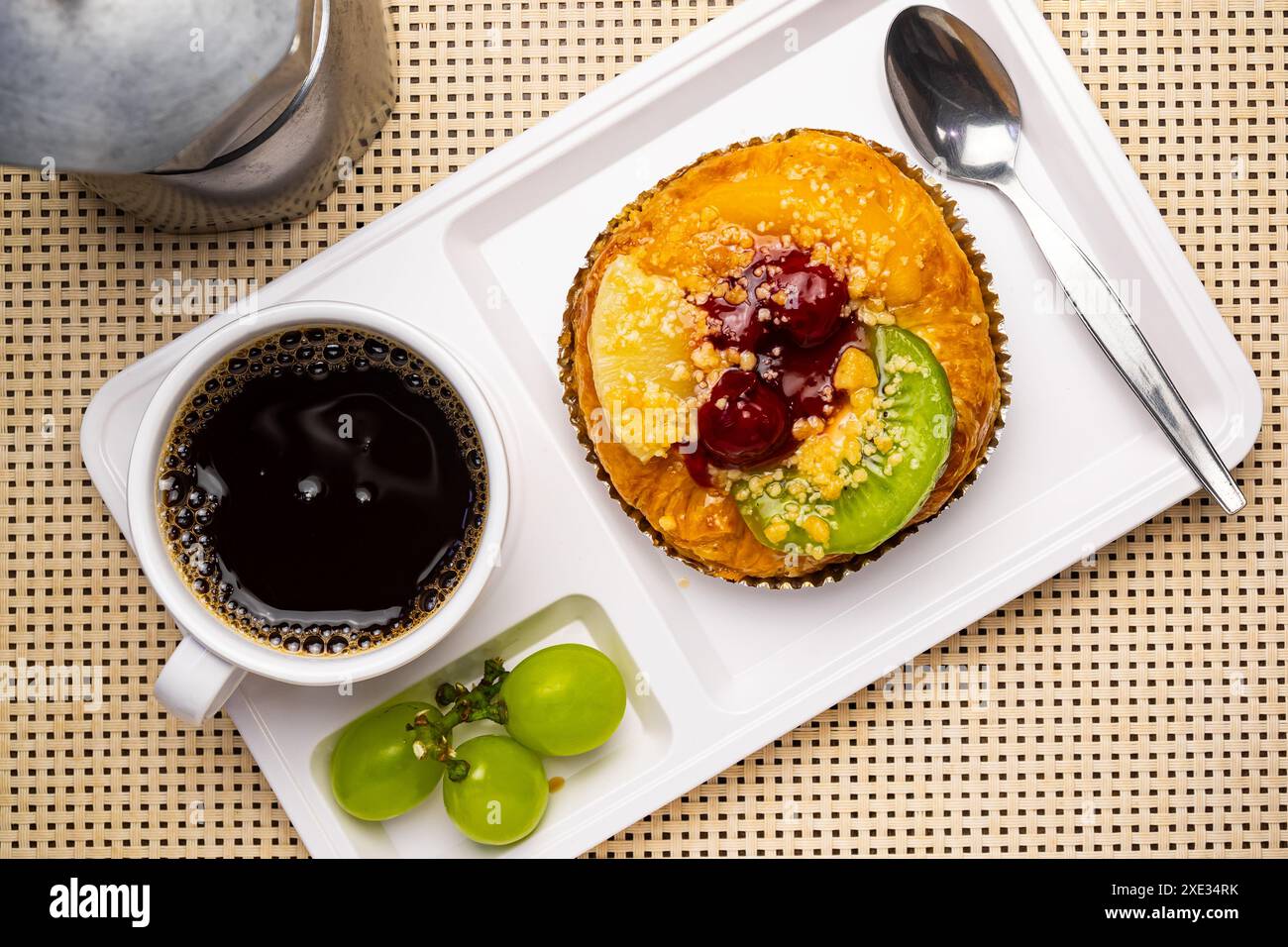 Top view of delicious homemade mixed fruit danish, a cup of black coffee, grapes and metal spoon in white plastic tray. Stock Photo