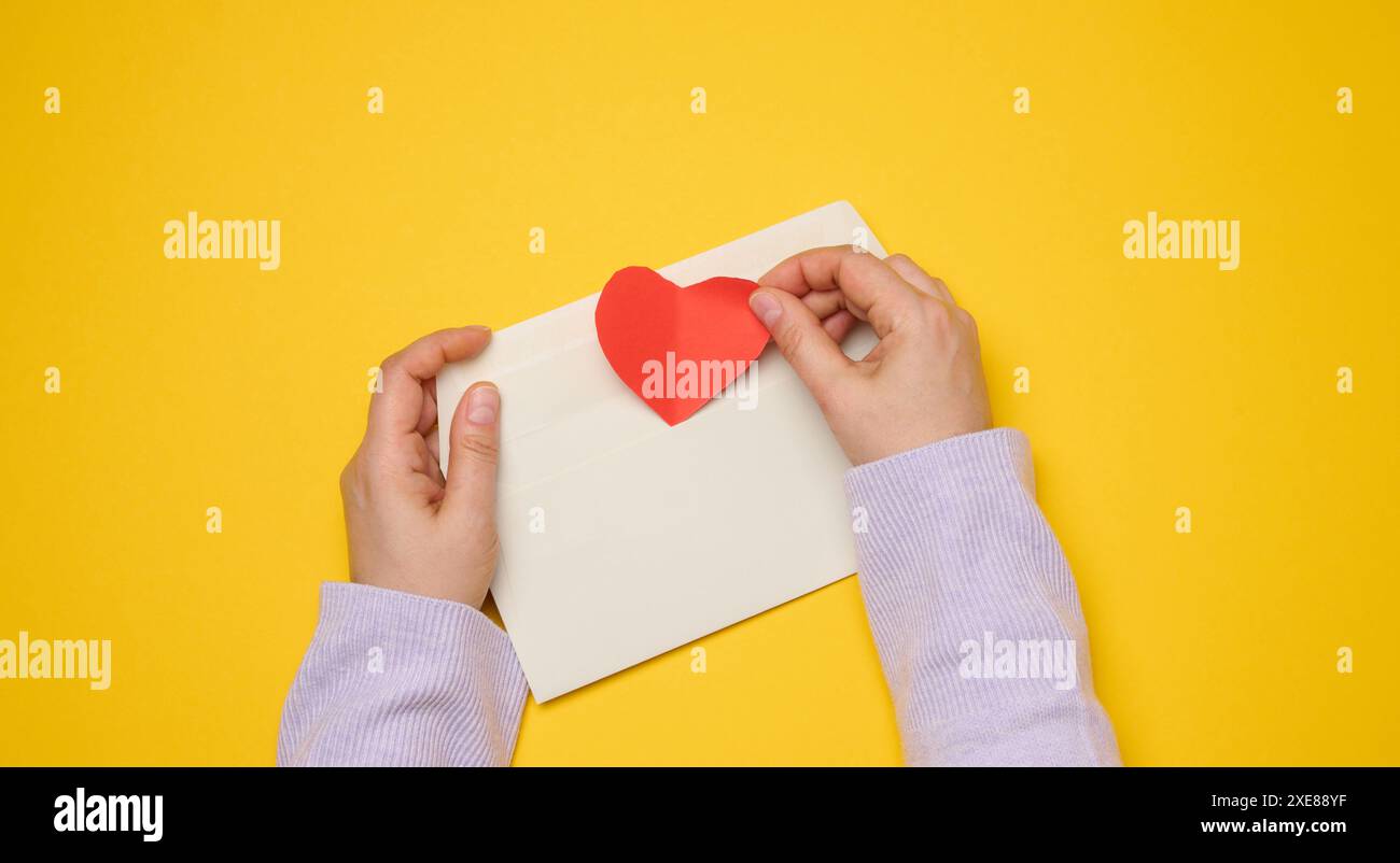 Female hands hold a white rectangular envelope on a yellow background, paper red hearts. Stock Photo
