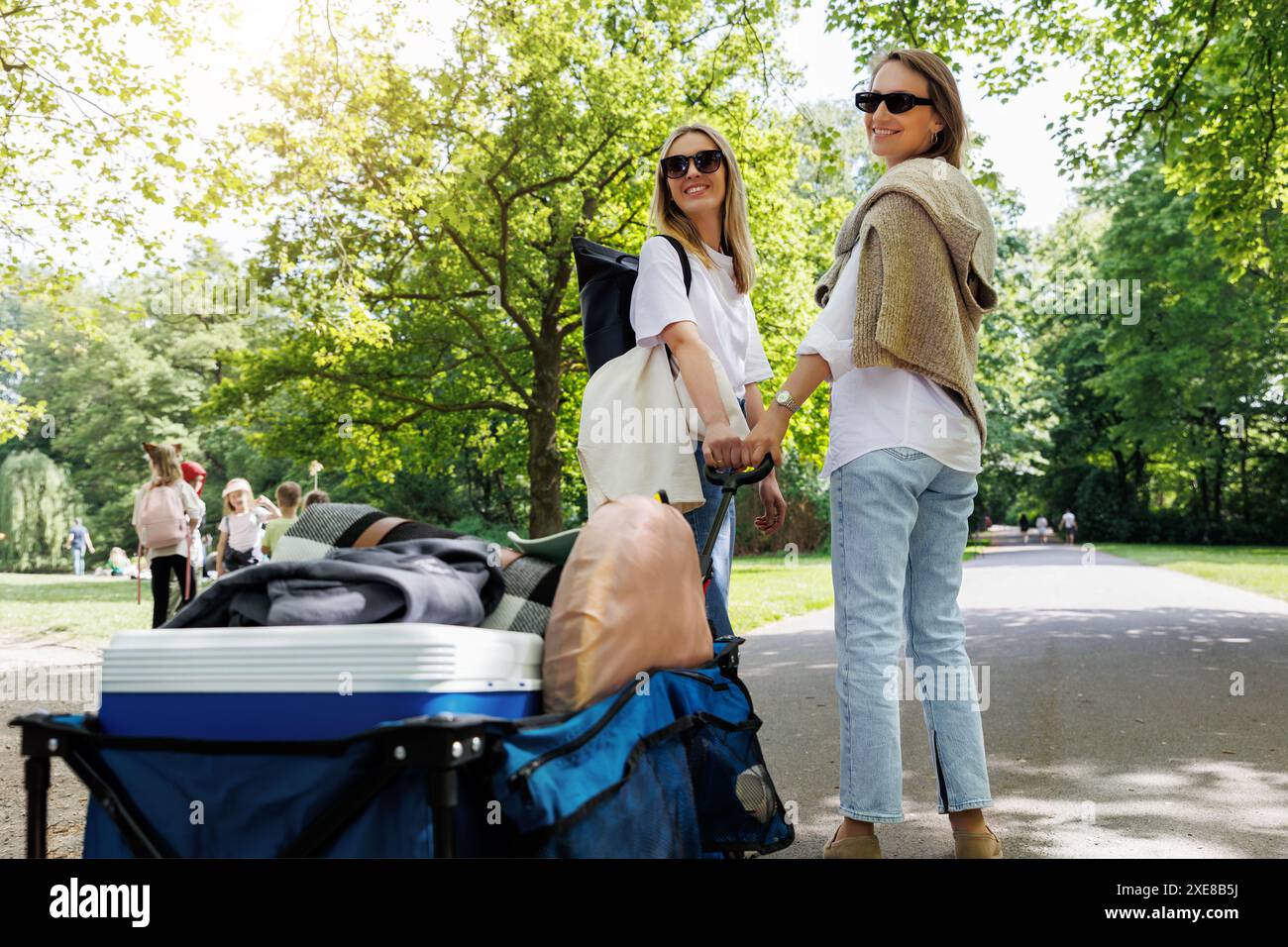 Back view two young adult beautiful woman girl friends enjoy have fun walking picnic party city park pulling cart food drink equipment sunny day Stock Photo