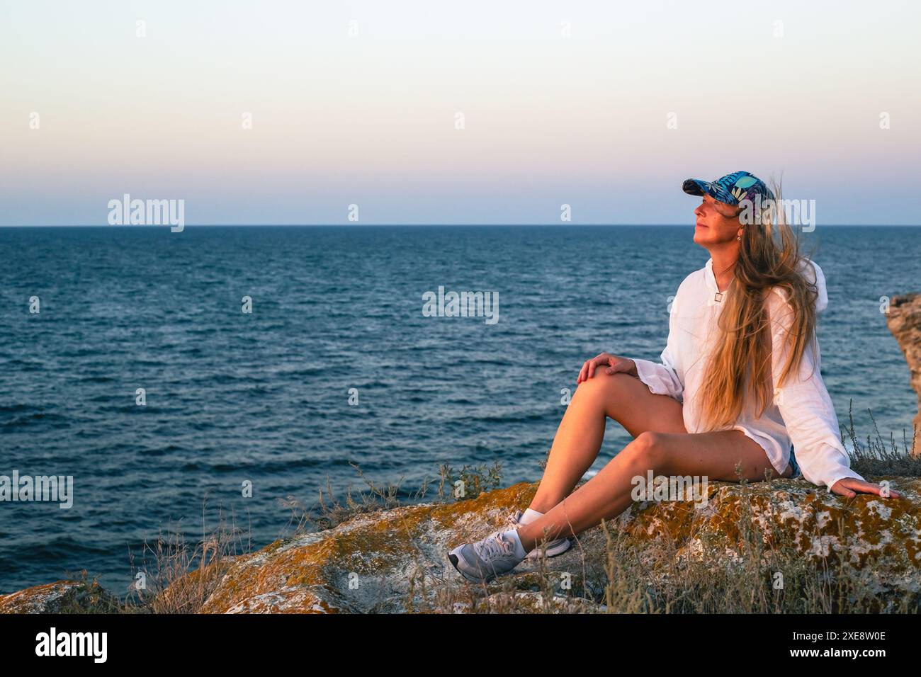 A woman with long hair sits on the rocks on the seashore at sunset ...