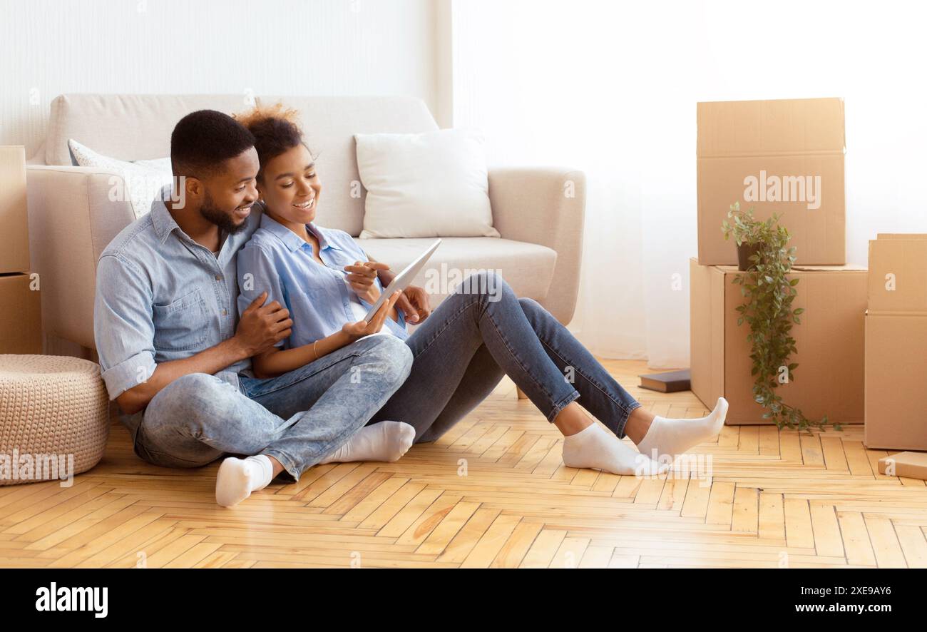 Spouses Using Tablet Sitting On Floor Among Unpacked Boxes Indoor Stock Photo