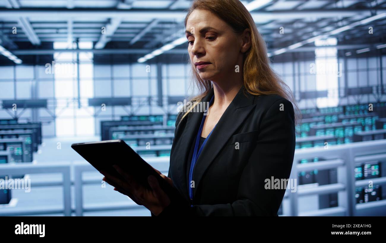Admin in server room checks recovery plan on tablet, monitoring equipment energy consumption. IT professional uses notebook in data center workplace, ensuring rigs are functioning optimally, camera B Stock Photo