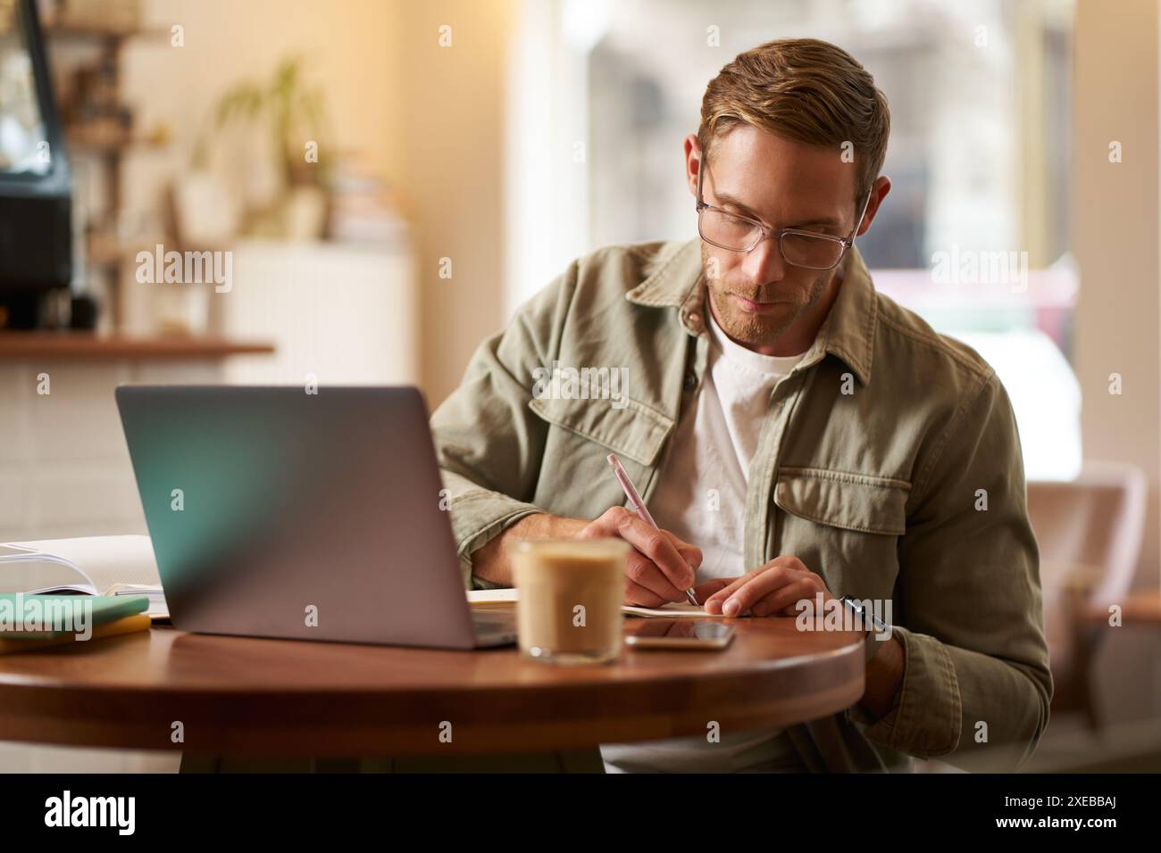 Image of focused young man in glasses, sitting in cafe, making notes, studying, attending online course, learning on remote from Stock Photo