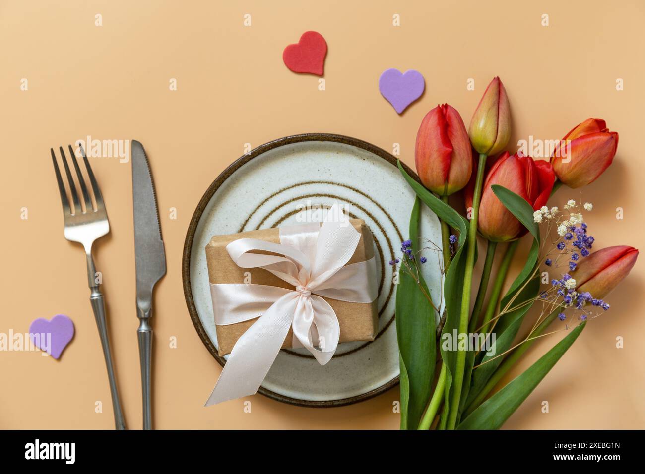 Beautiful table setting with gift box, decorative hearts and tulips flowers on pastel peach background. Stock Photo