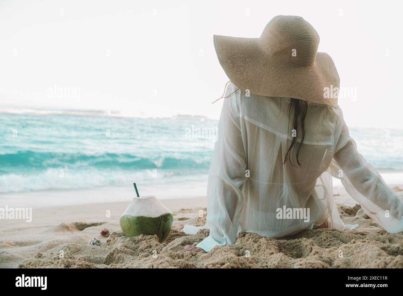 Back view of a young woman in a straw hat sits on the beach and drink ...